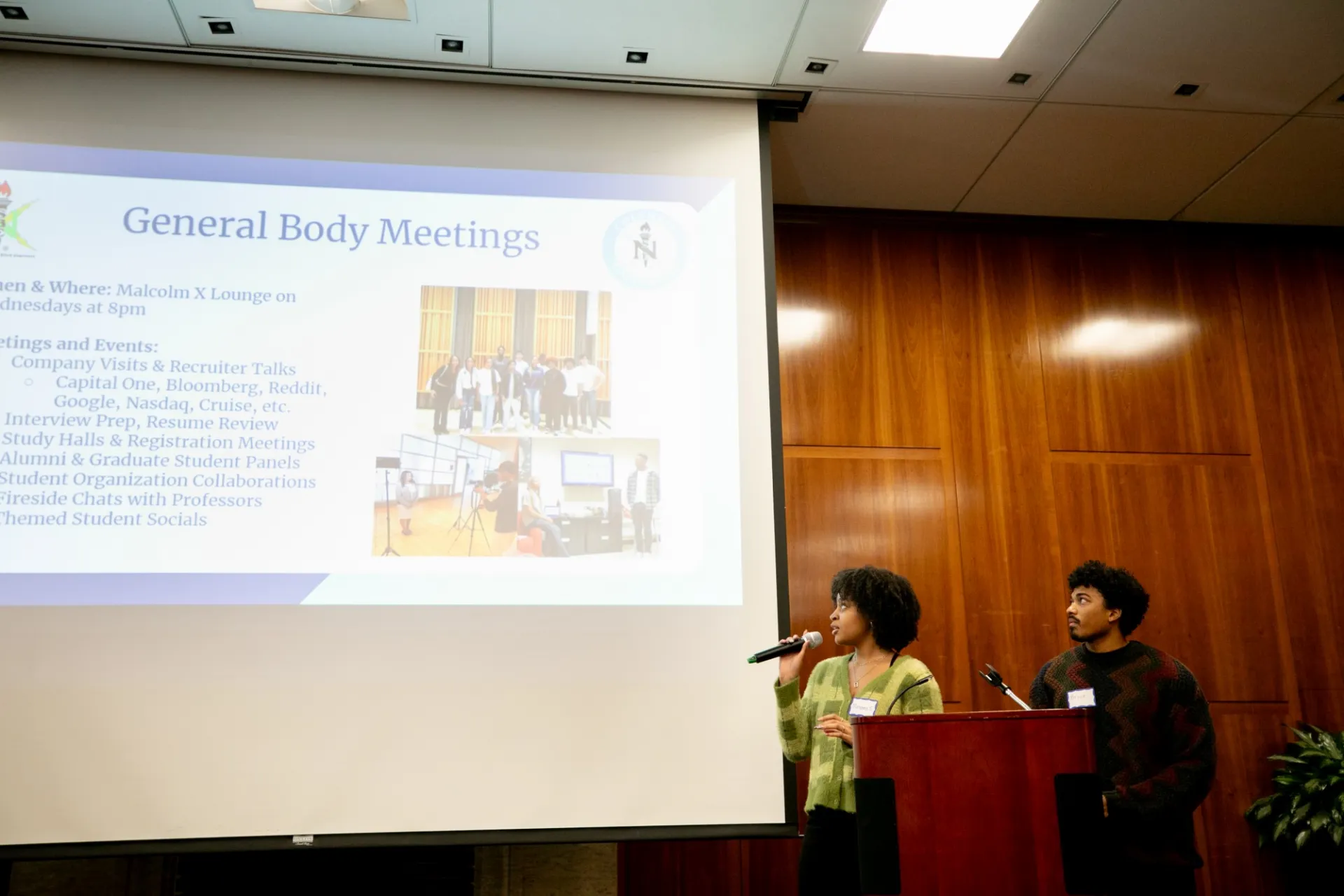 Speakers presenting to an audience. The presentation slide next them is titled "General Body Meetings".