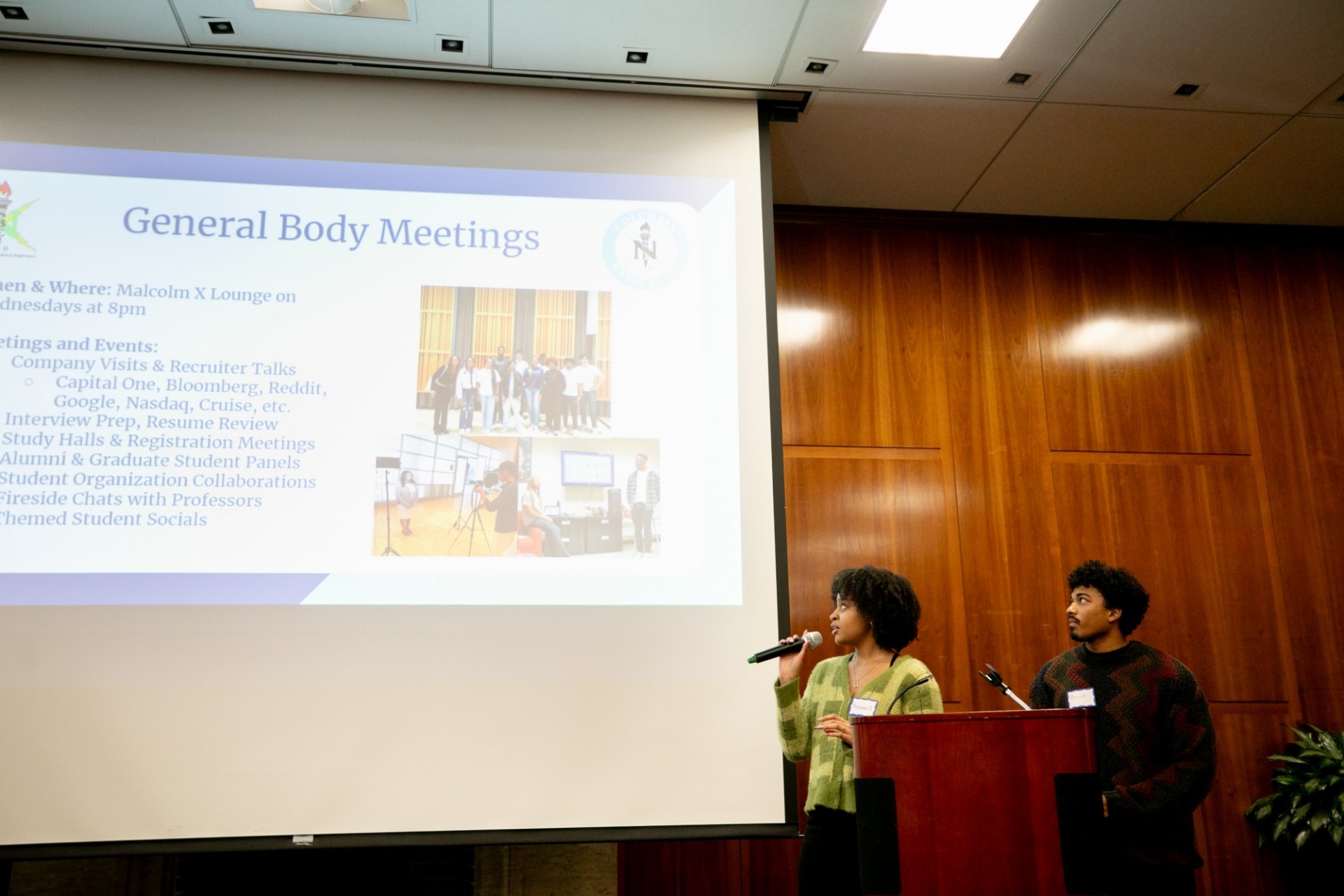 Speakers presenting to an audience. The presentation slide next them is titled "General Body Meetings".