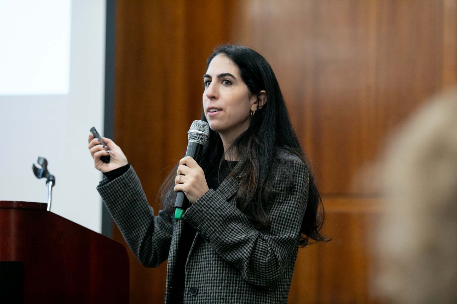 A speaker holding a microphone and clicker addresses an audience.