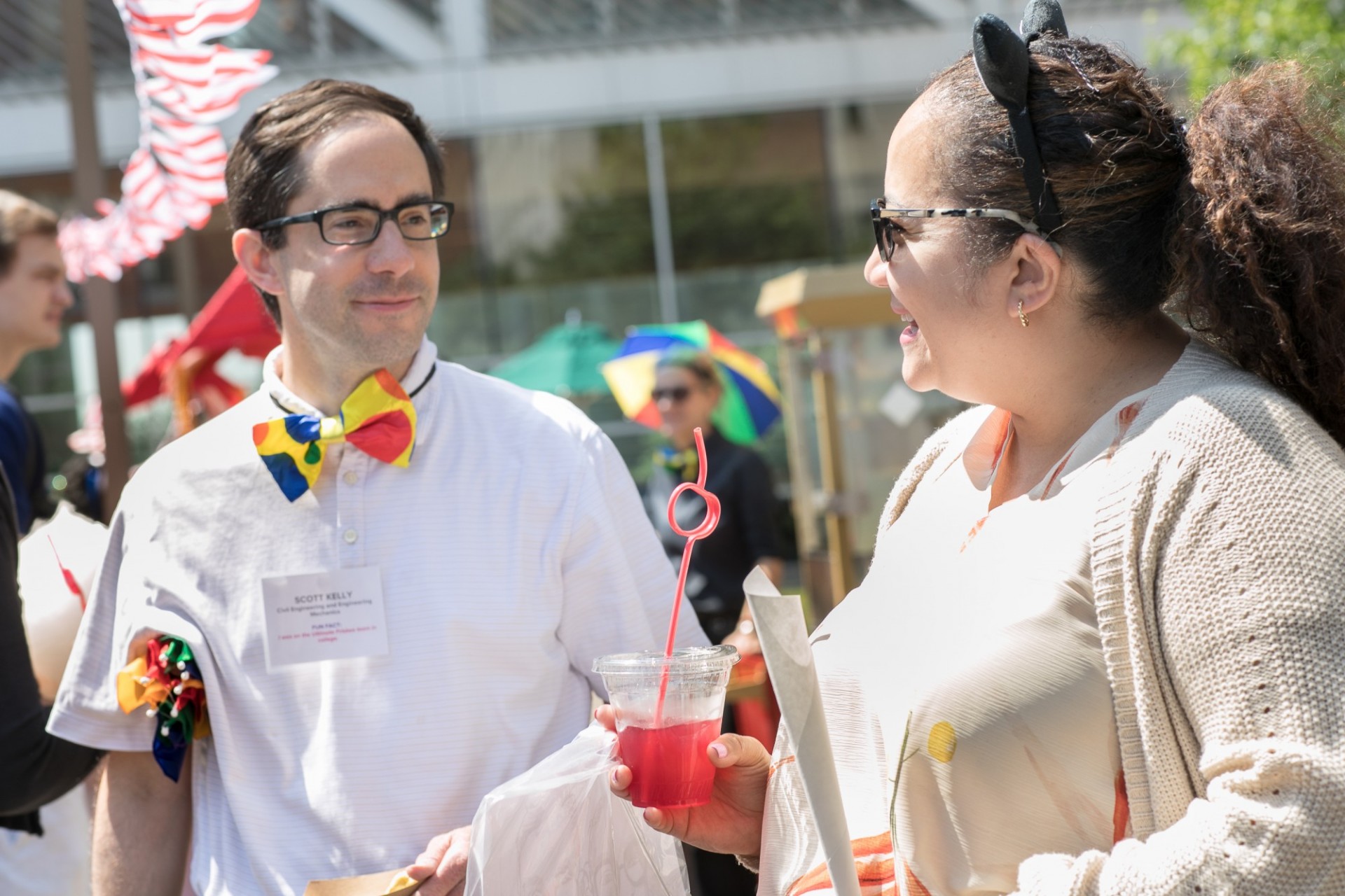 Staff chatting and enjoying refreshments wearing carnival themed carnival accessories. One is wearing a clown bowtie and the other is wearing bat ears.