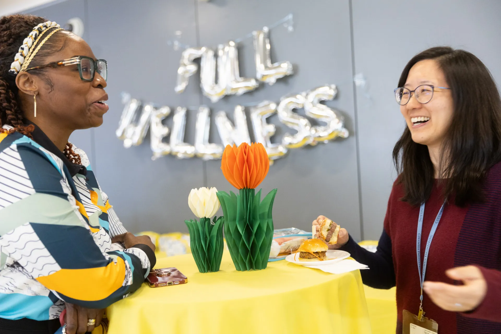 Two staff members chatting with two flowers in the middle and full wellness banner in the background 